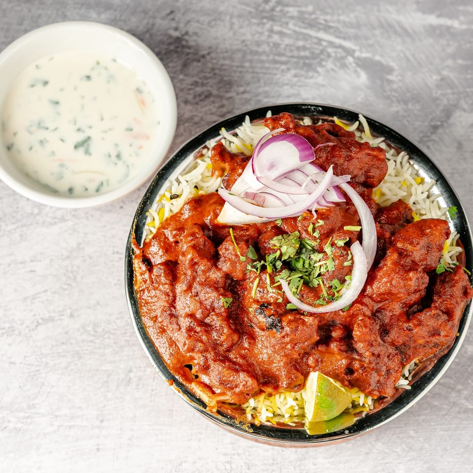 Indian-style spiced chicken over biryani rice, garnished with red onion, cilantro, and lime, served with creamy herb sauce.