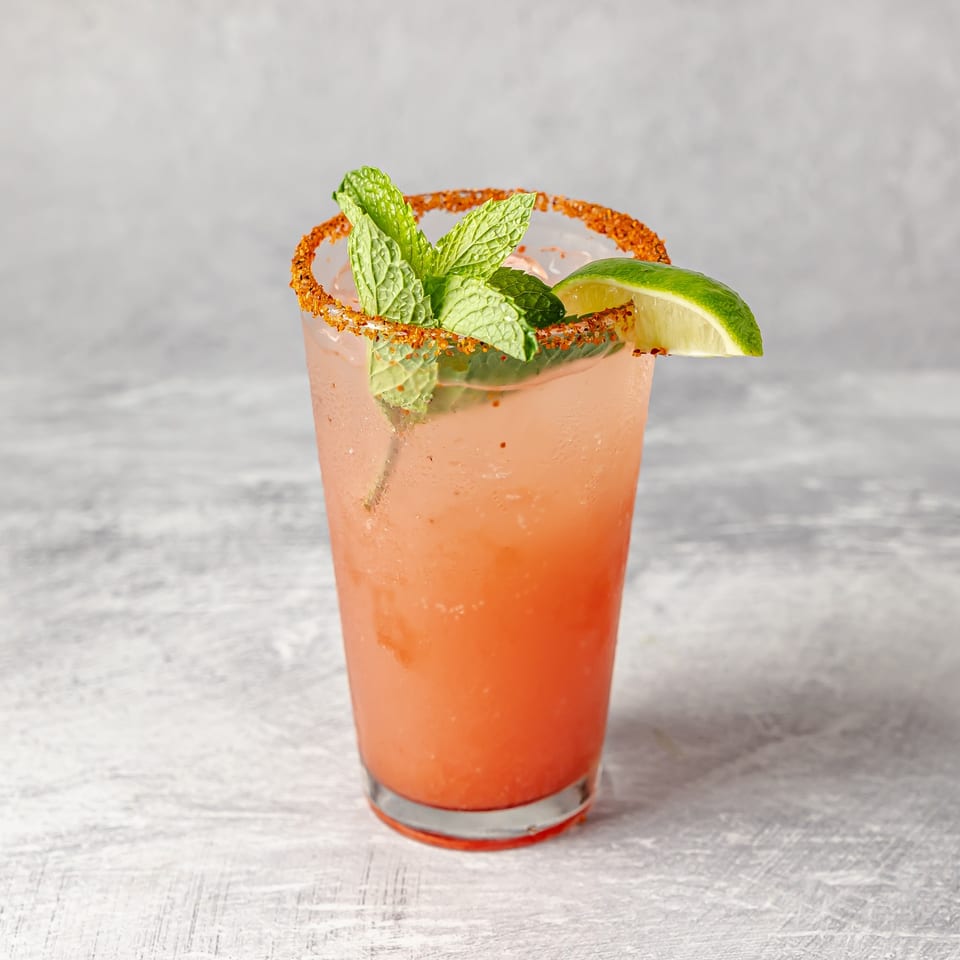 Refreshing pink cocktail with a chili-salt rim, mint leaves, and lime wedge in a glass on a tabletop.