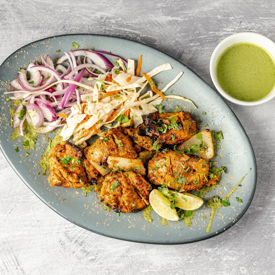 Grilled spiced chicken with onions, coleslaw, cilantro, lime wedges, and green chutney on a platter.