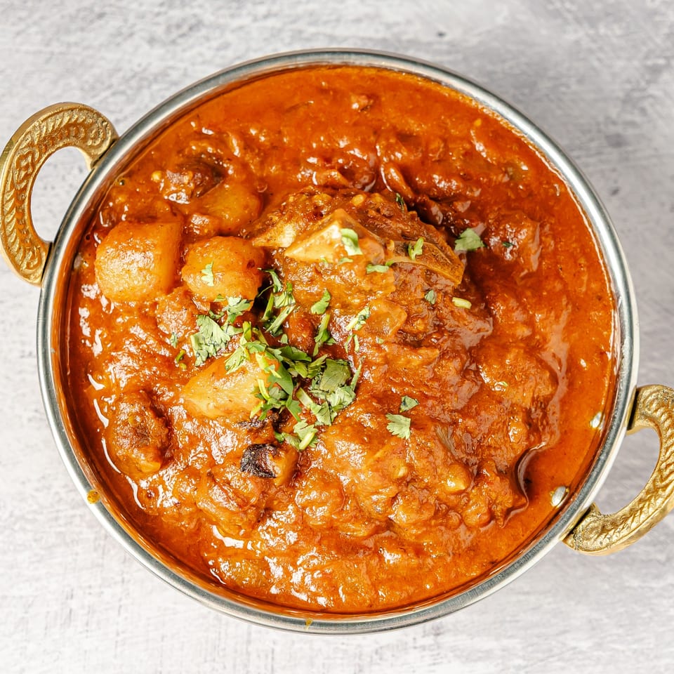 Overhead view of an Indian curry with potatoes and meat in rich red sauce, garnished with fresh cilantro in a handled serving bowl.