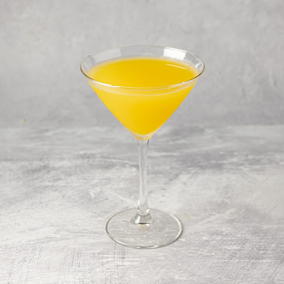 Bright yellow cocktail served in a clear martini glass on a light gray tabletop.