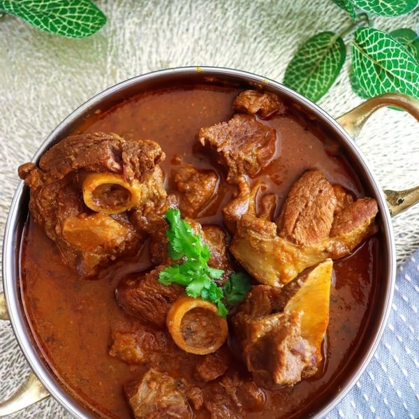 Close-up of a metal bowl filled with rich, reddish-brown lamb curry garnished with fresh cilantro.