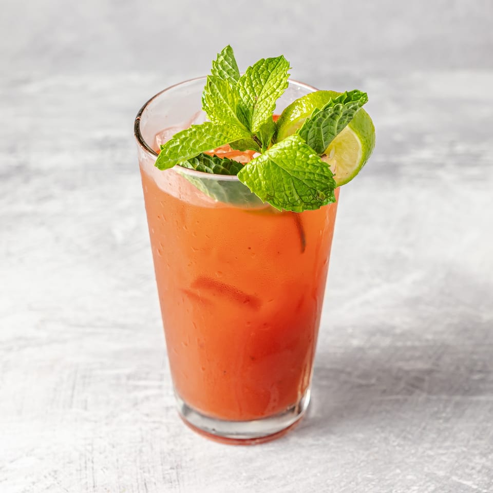 Refreshing orange cocktail in a glass, garnished with fresh mint leaves and lime wedges on a light tabletop.