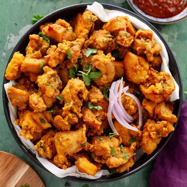 A black bowl filled with golden-brown fried chicken pakora, garnished with red onion and cilantro.