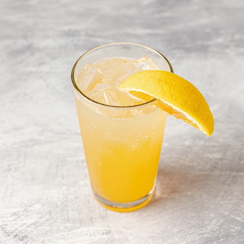 Chilled yellow citrus cocktail with ice in a clear glass, garnished with a large lemon peel on a marble tabletop.