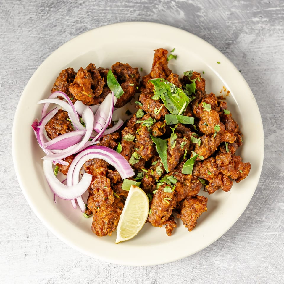 Plate of crispy spiced fried chicken garnished with curry leaves, cilantro, sliced red onion, and a lime wedge.