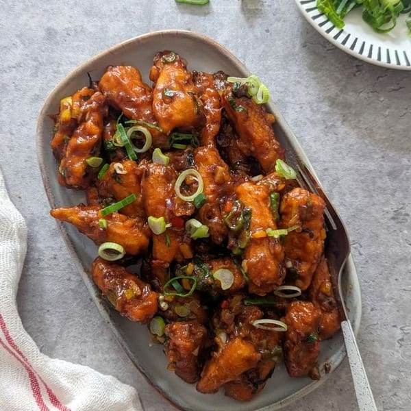 Crispy chili paneer, coated in a rich sauce and garnished with fresh green onions, on a gray plate.